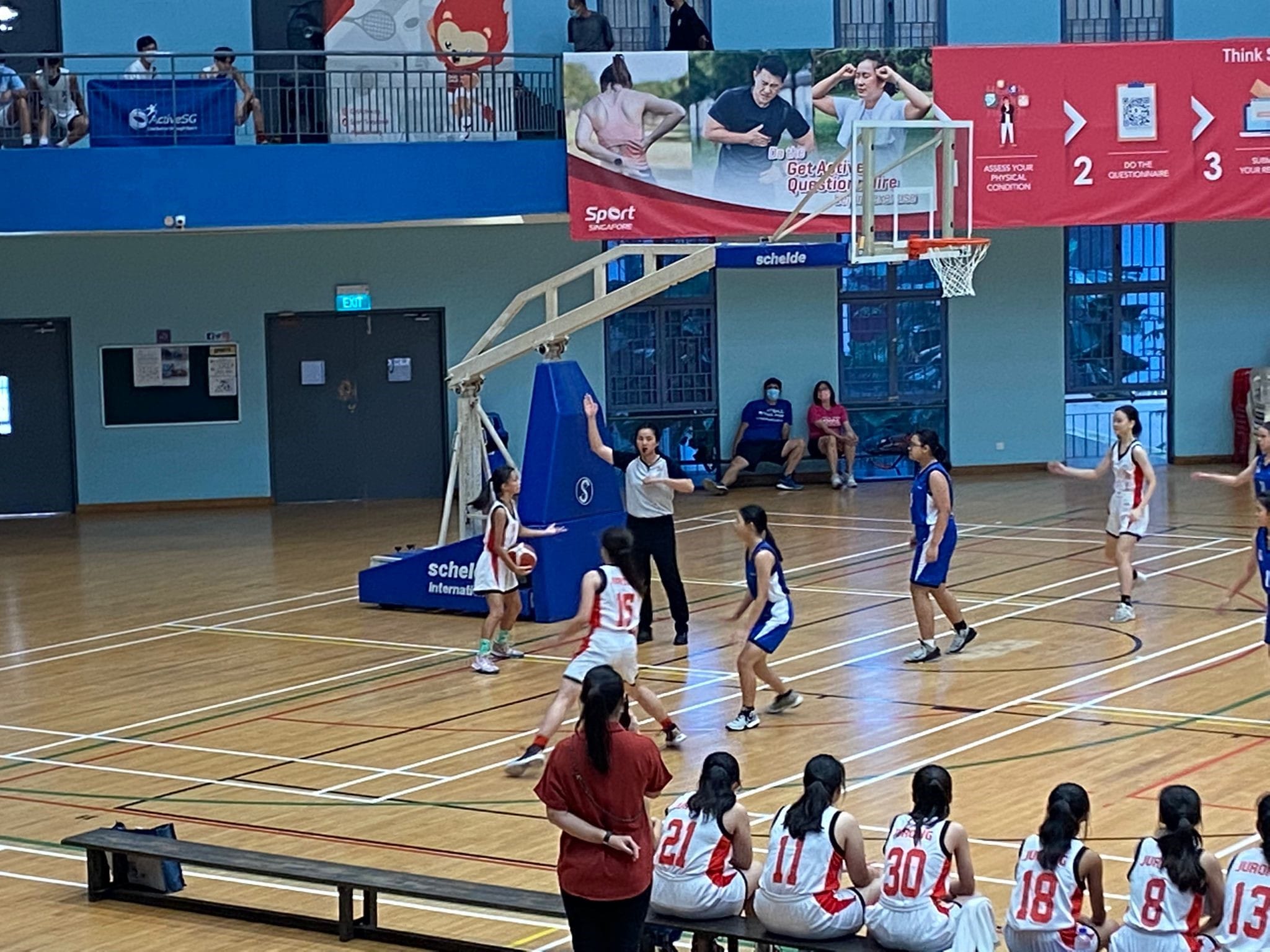 Girls in white and blue uniforms play basketball indoors, with a referee and seated substitutes watching nearby.